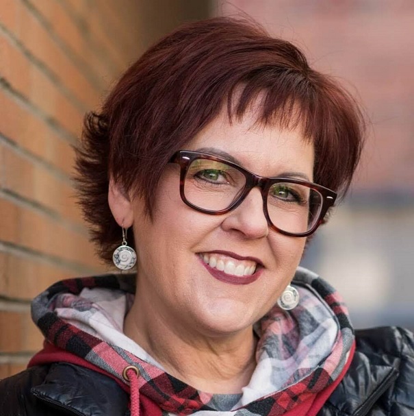 A woman with short auburn hair and dark-framed glasses smiling in front of a brick wall.