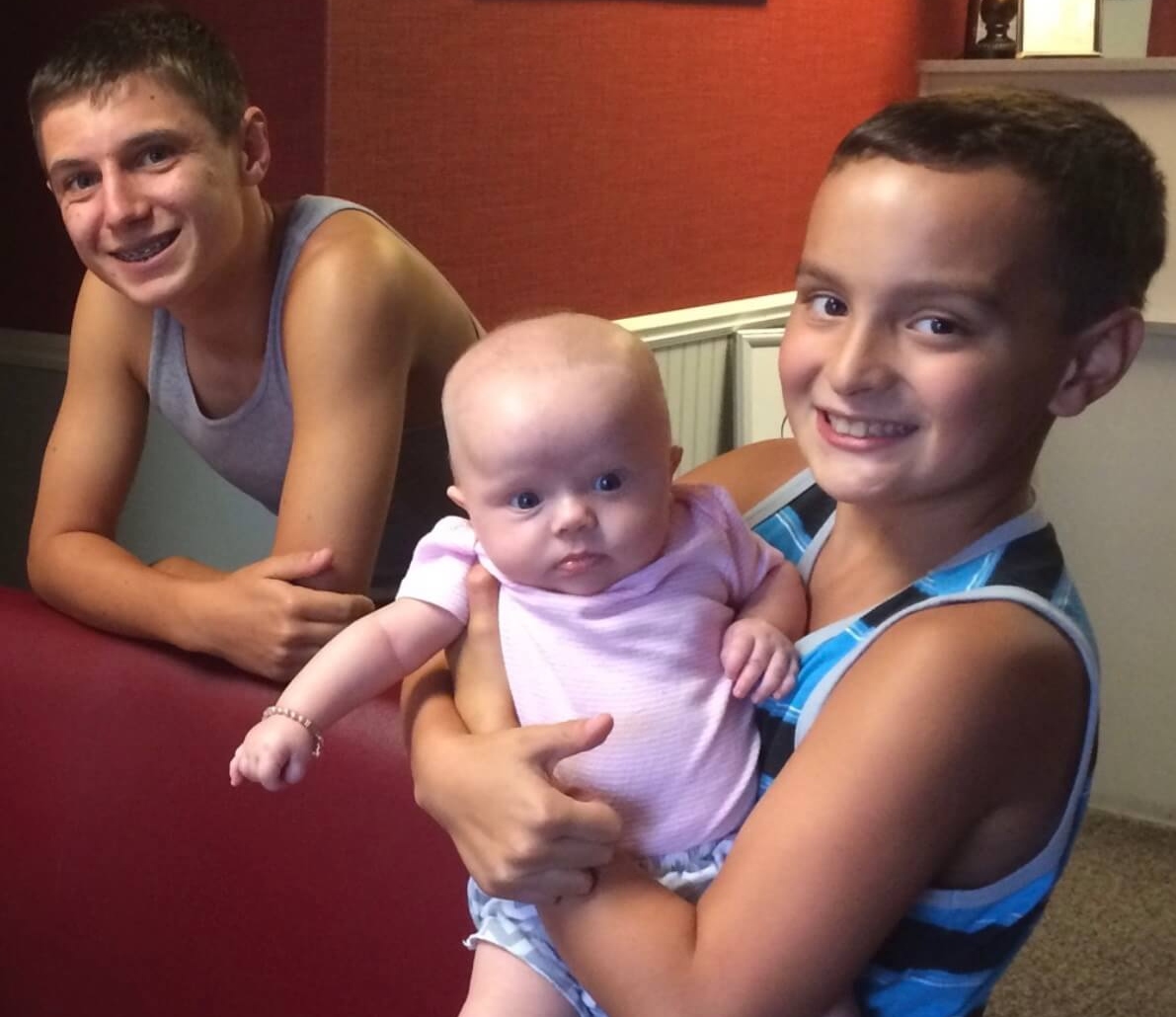 Two teenage boys in vests standing together, one of them holding a baby in a pink bodysuit.