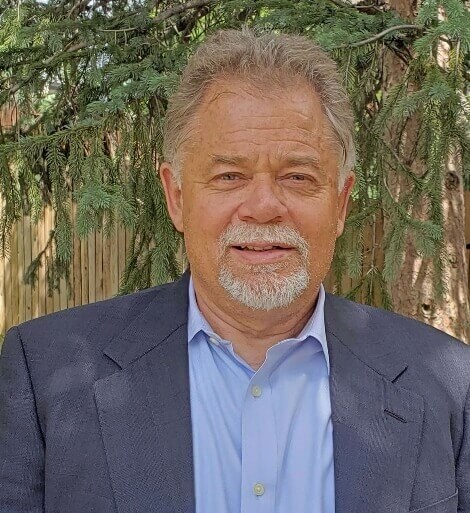A man with grey hair and a beard in a blazer and open-necked shirt, smiling in front of an evergreen tree.