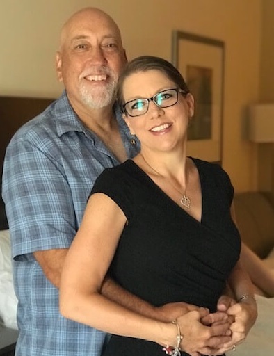 A man and a woman standing together indoors with their arms around each other, smiling.