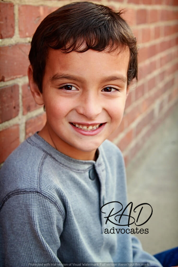 A smiling boy in a grey long-sleeved top standing in front of a brick wall.