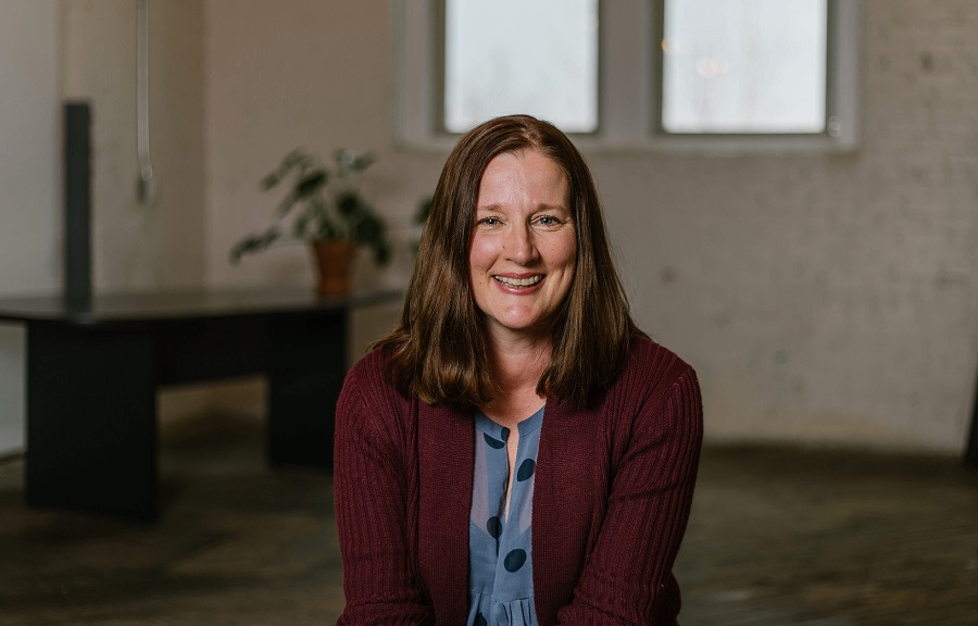 A woman in a dark red cardigan smiling, seated in a bright room with a plant behind her.