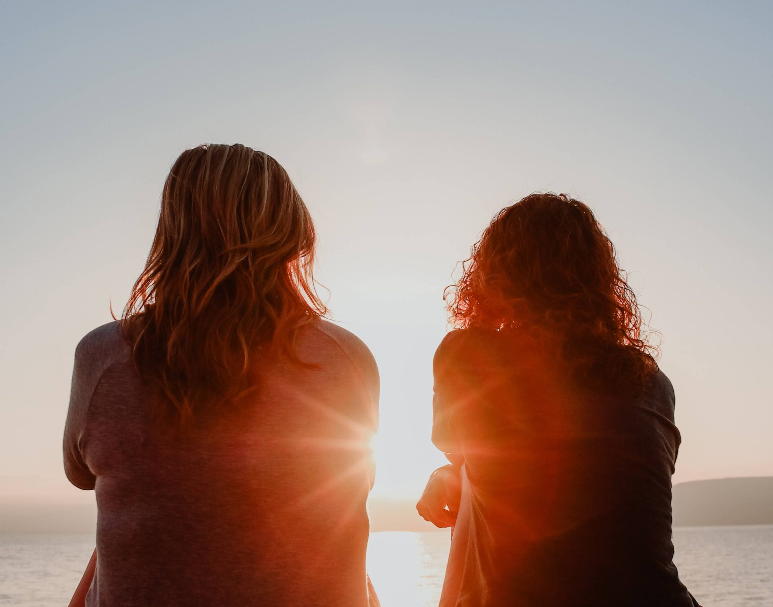 Two women sitting side by side on a hillside at sunset, backs to the camera, looking out over water.