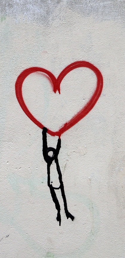 Street art on a concrete wall: a small stick figure hanging from a red spray-painted heart.