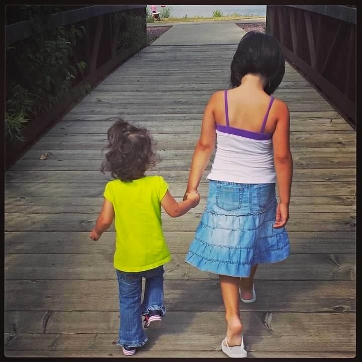 Two girls seen from behind, holding hands as they walk across a covered wooden bridge.