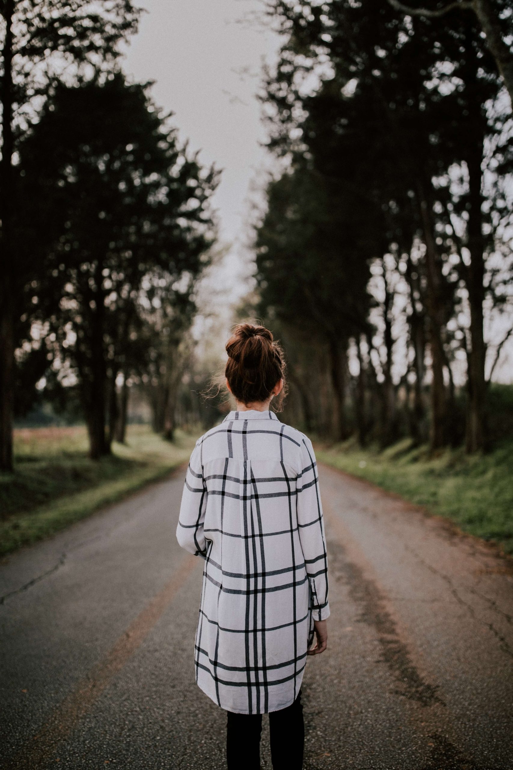 A woman in a checked coat seen from behind, walking down a tree-lined road.