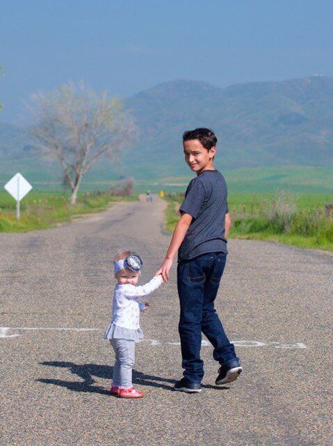 A boy walking along a country road holding the hand of a toddler in a white coat, hills behind them.