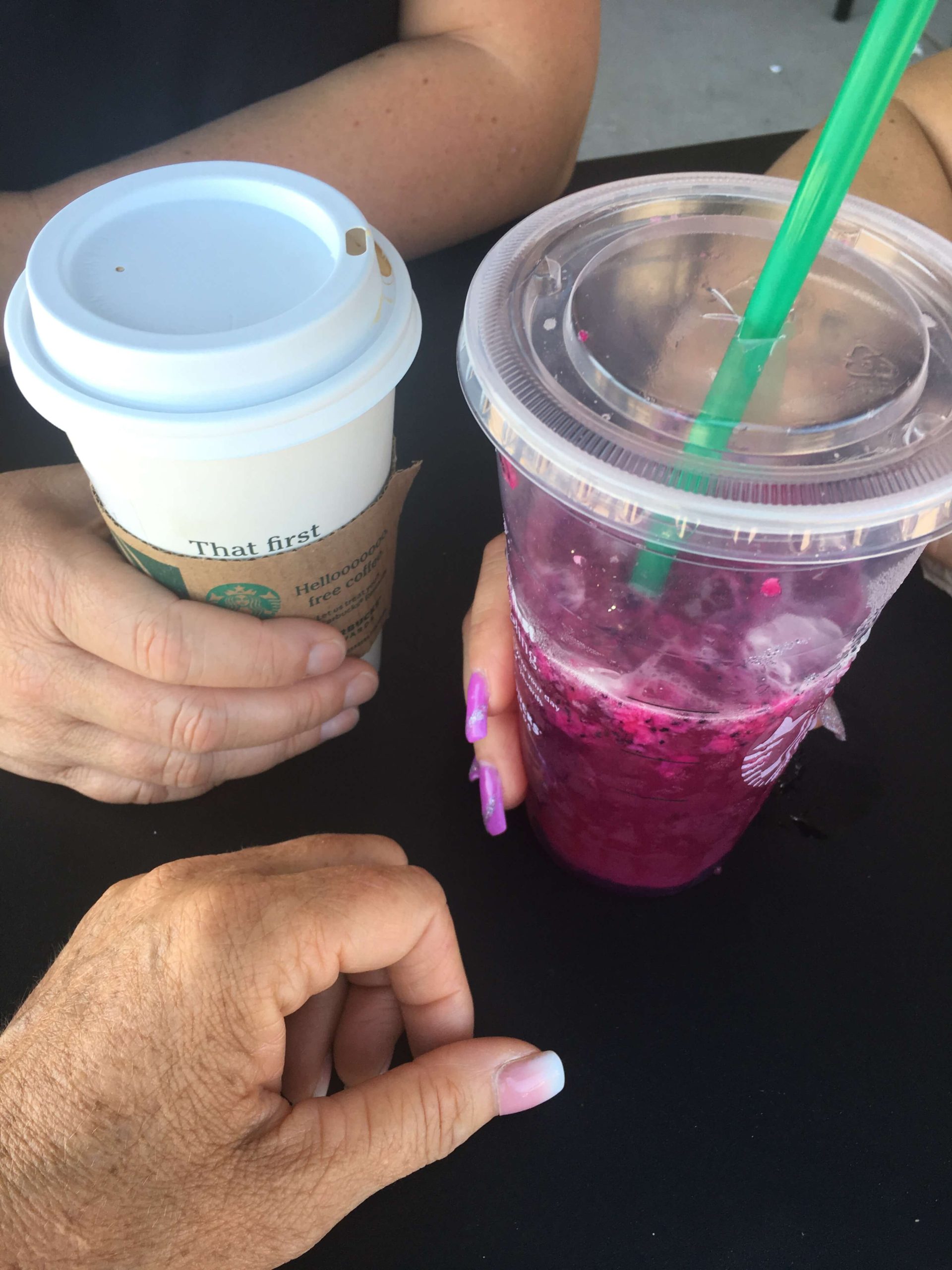 Two people sitting at a table holding drinks — a takeaway coffee cup and an iced pink drink.