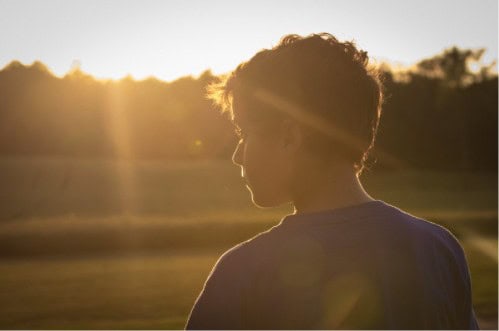 A young person silhouetted against a low sun over open fields.