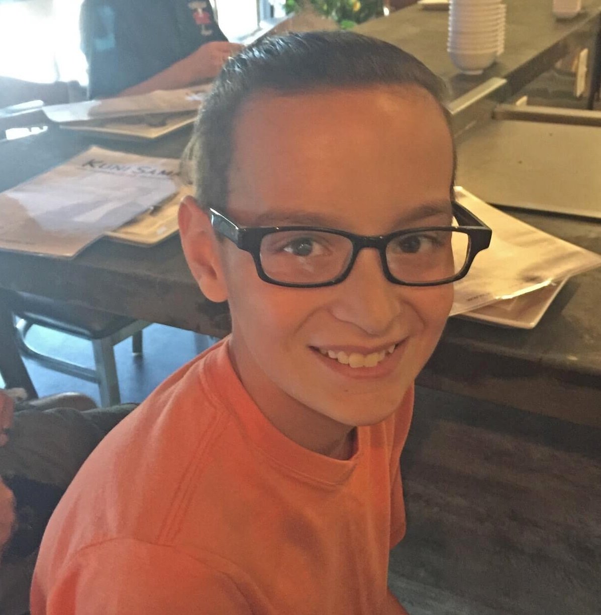 A boy in glasses and an orange top smiling at the camera in a restaurant.