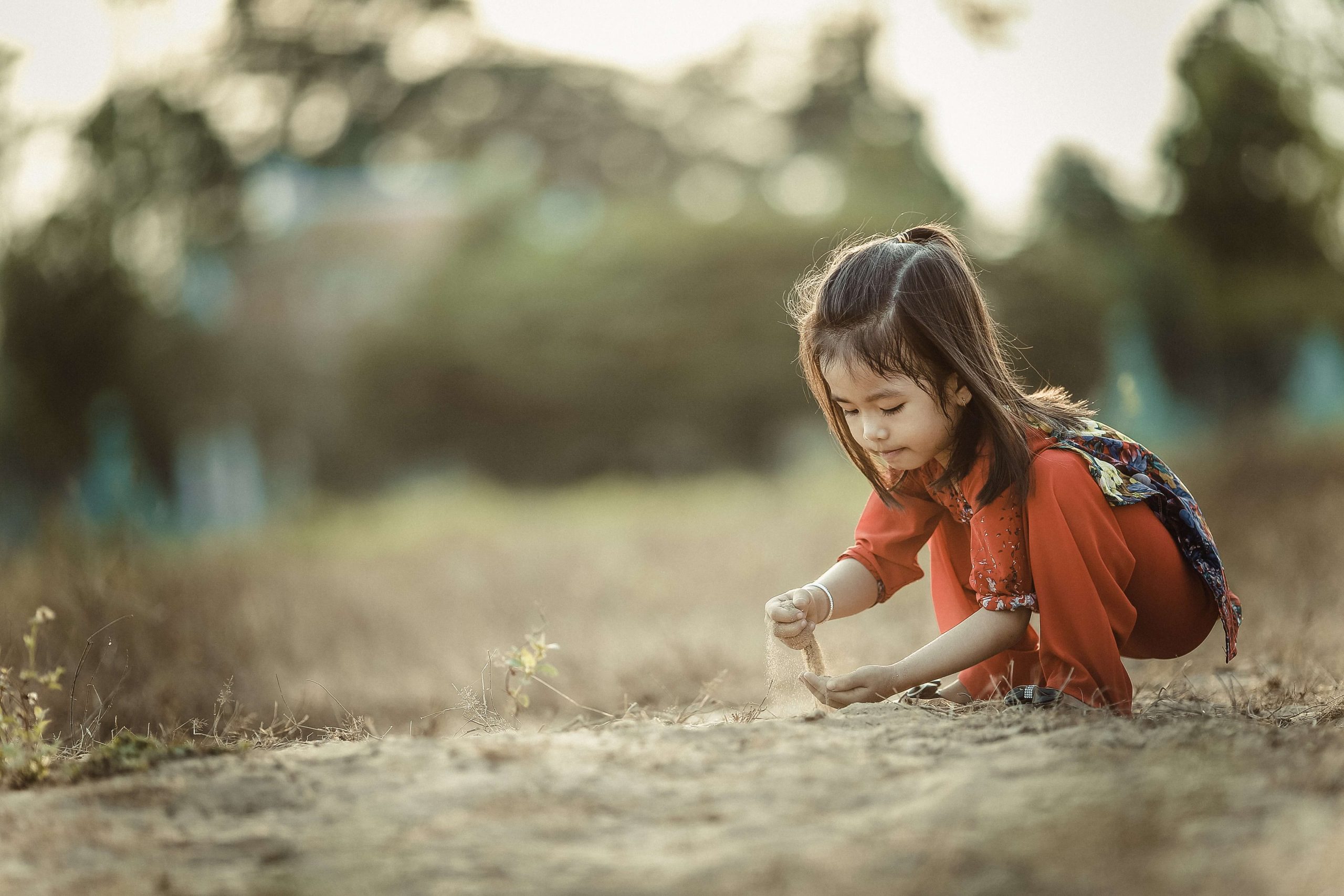 A young girl in an orange top crouching alone in a field, letting sand run through her fingers.