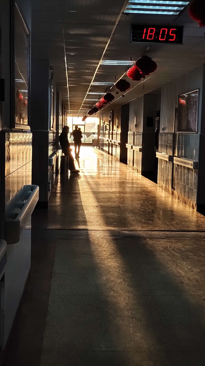 A long institutional corridor lit by low sun, with a digital clock reading 18:05 overhead.