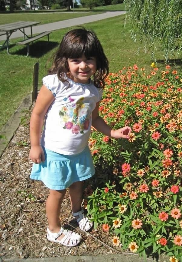 A young girl in a white top and blue skirt standing beside a bed of orange and red flowers.