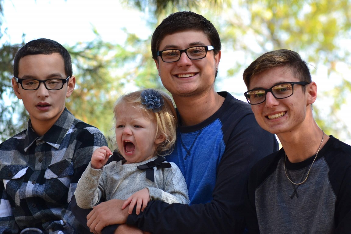 Three teenage boys standing together outdoors, one holding a small child who is mid-yawn.