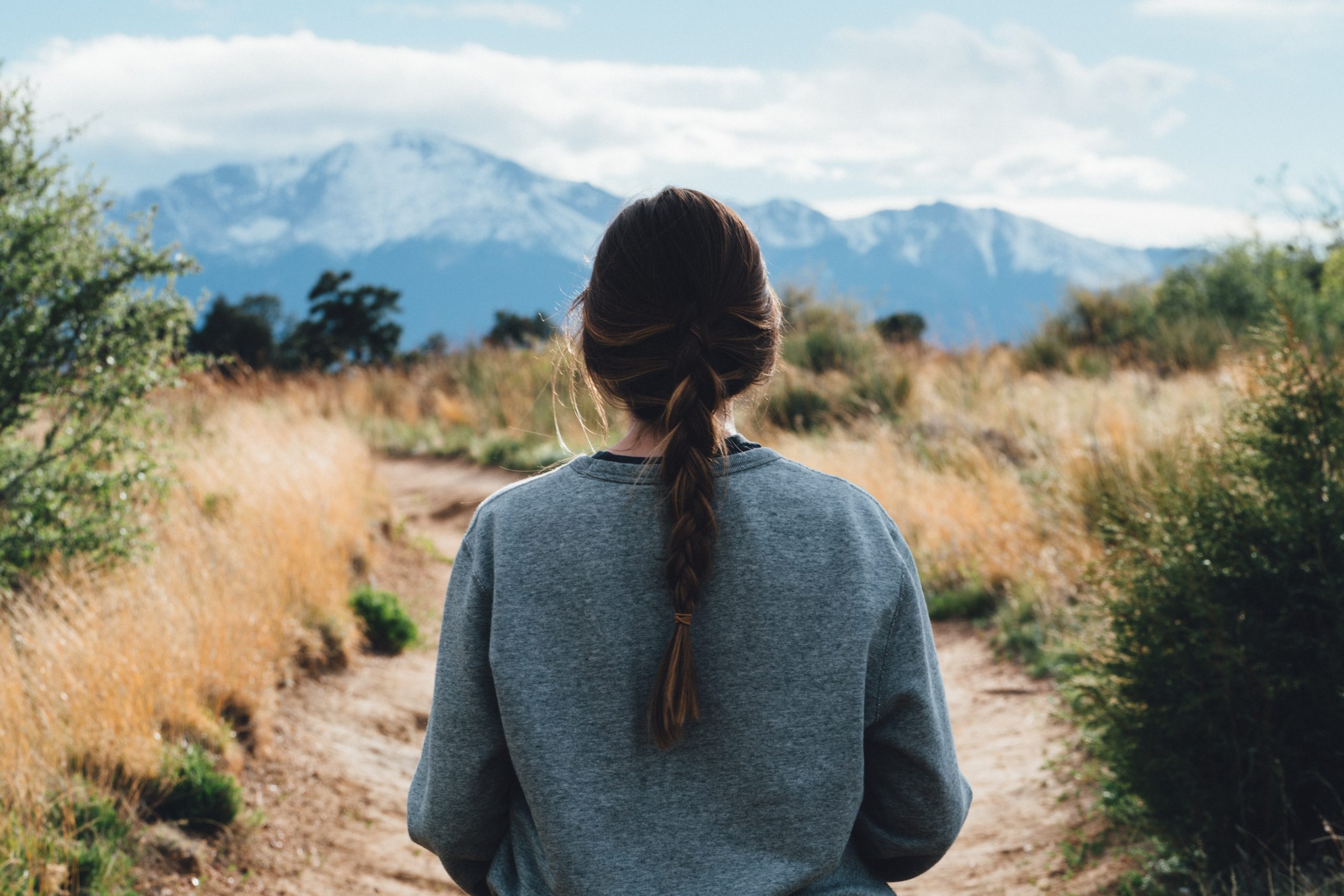 A woman with a plaited ponytail, seen from behind, walking a dirt path towards distant mountains.