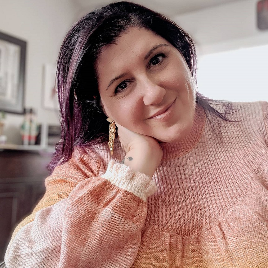 A woman with dark purple-tinted hair in a pink jumper, resting her chin on her hand and smiling.