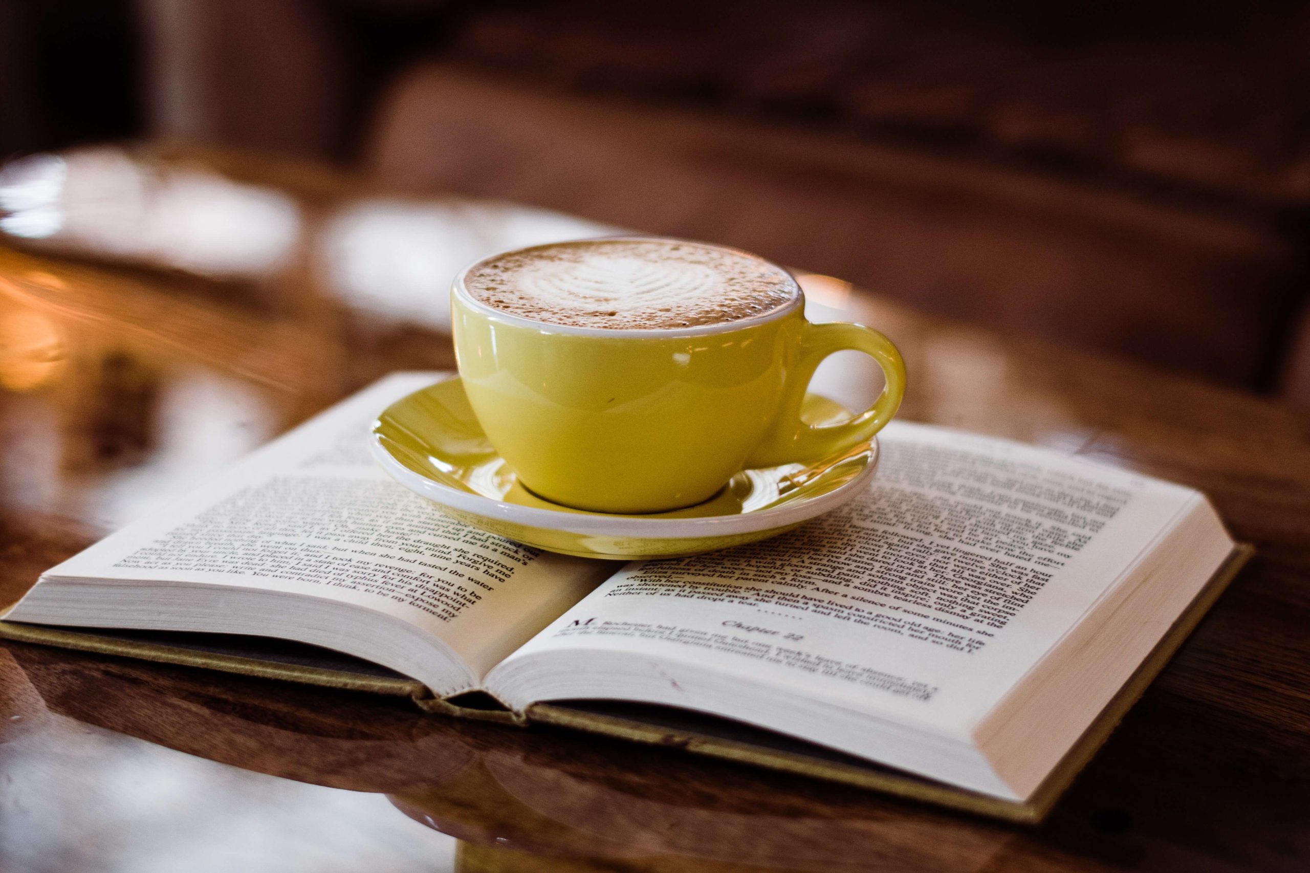 A yellow cup of coffee resting on an open book on a wooden table.
