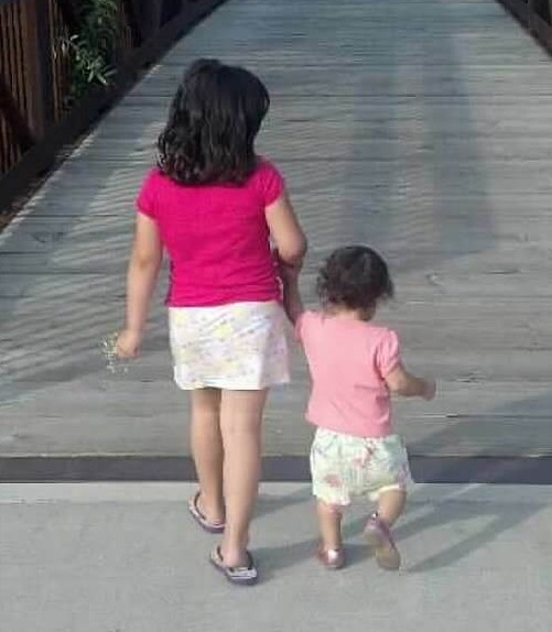 Two girls walking away along a wooden footbridge, the older one holding the younger's hand.