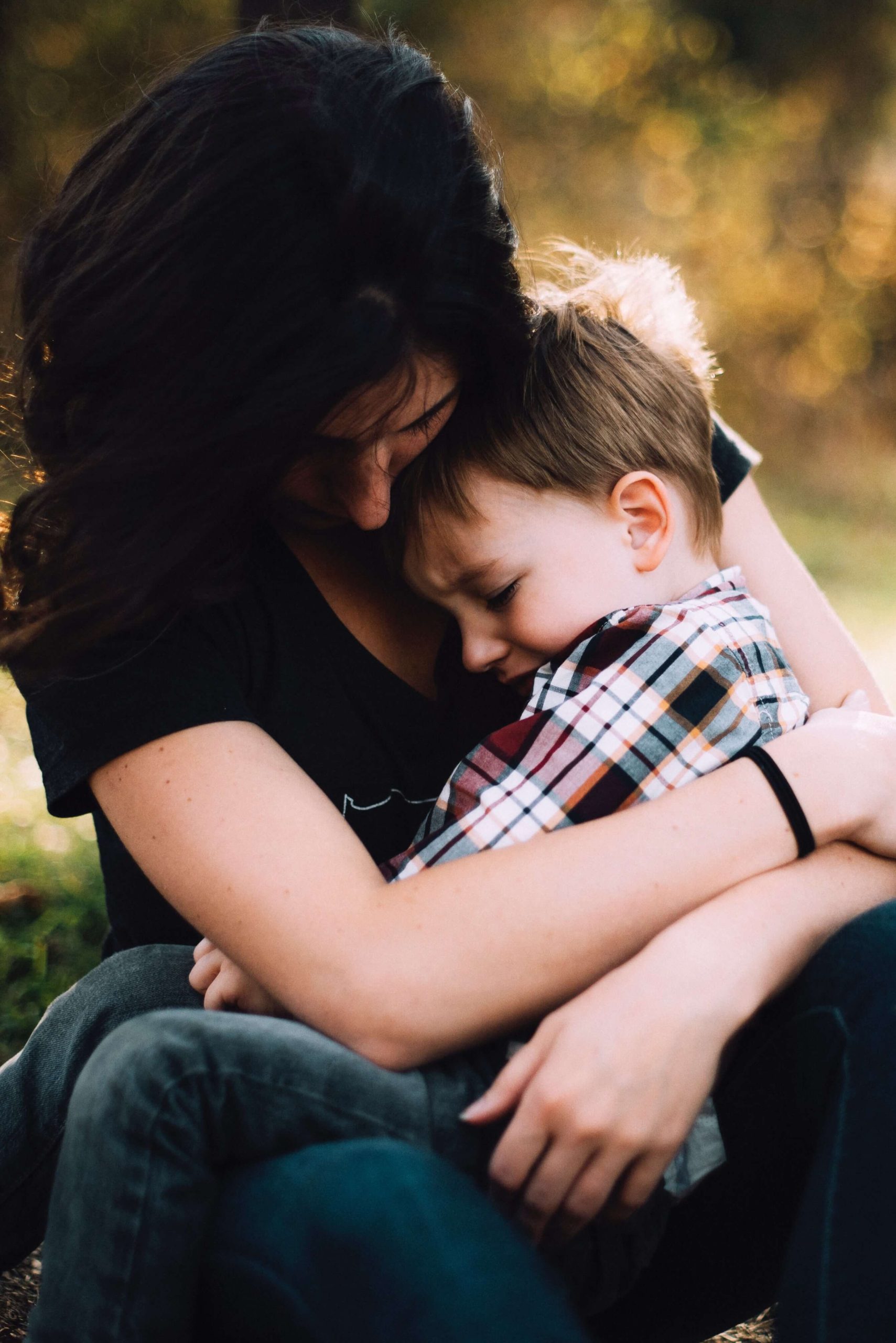A woman sitting outdoors holding a young boy in her lap, her cheek resting against his head.