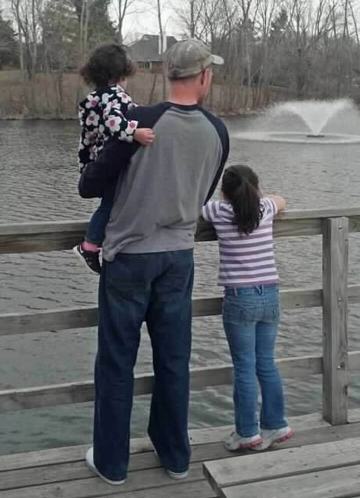 A man in a baseball cap at a lakeside railing with a small child on his hip and an older girl beside him, a fountain behind them.