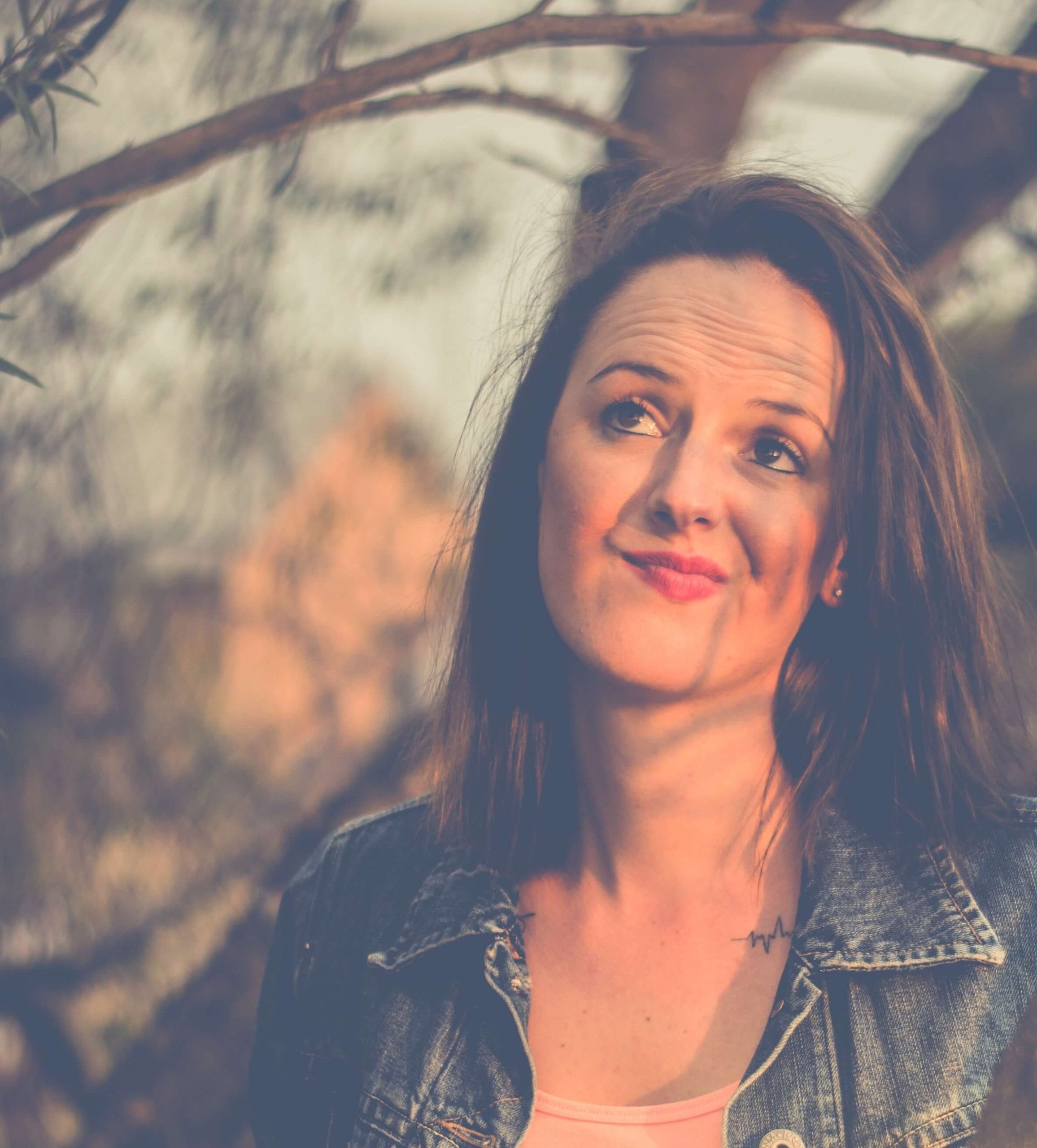A woman in a denim jacket leaning against a tree, looking upward with a wry half-smile.