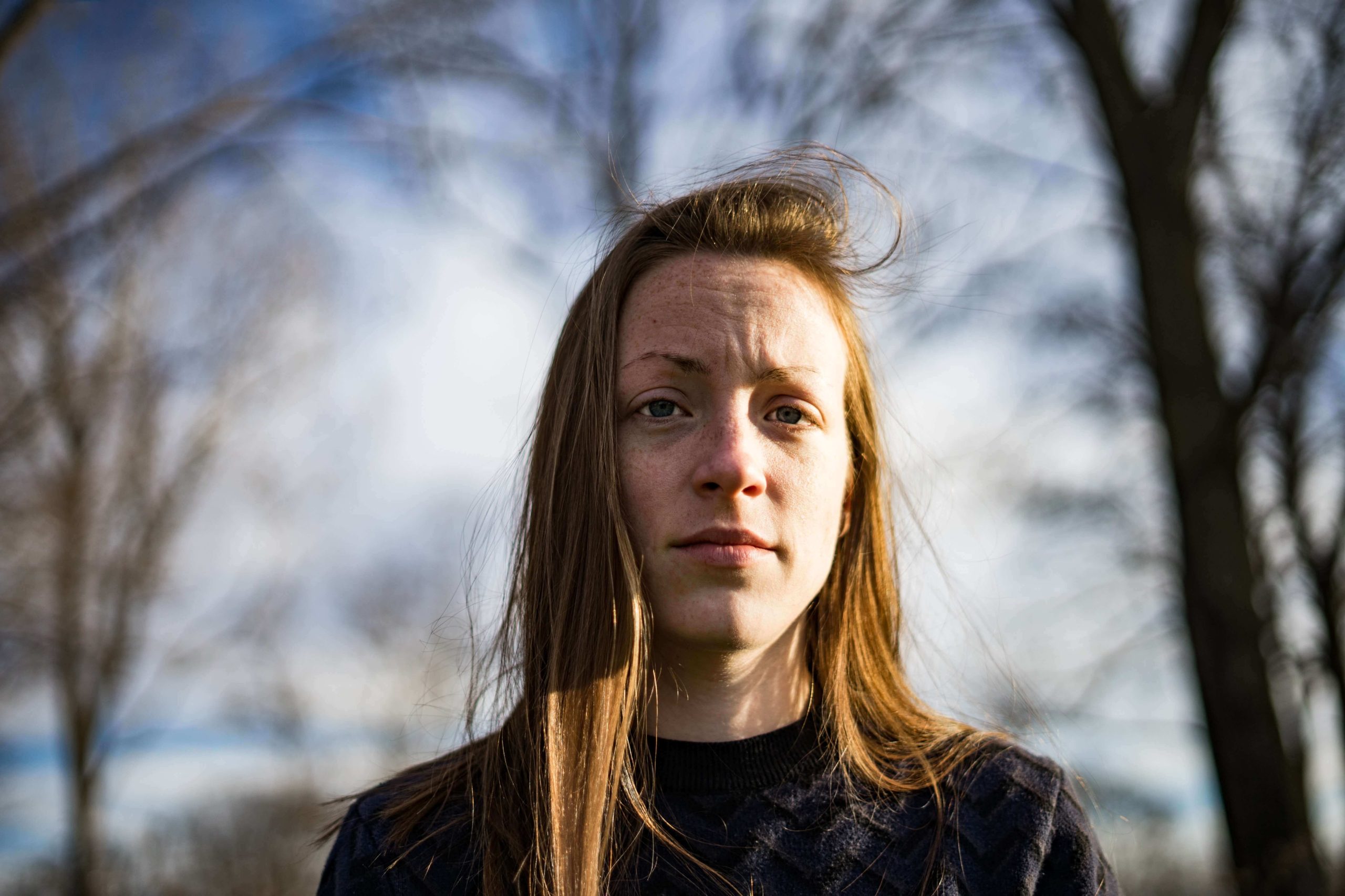 A woman with long hair standing outdoors among bare winter trees, looking directly at the camera.