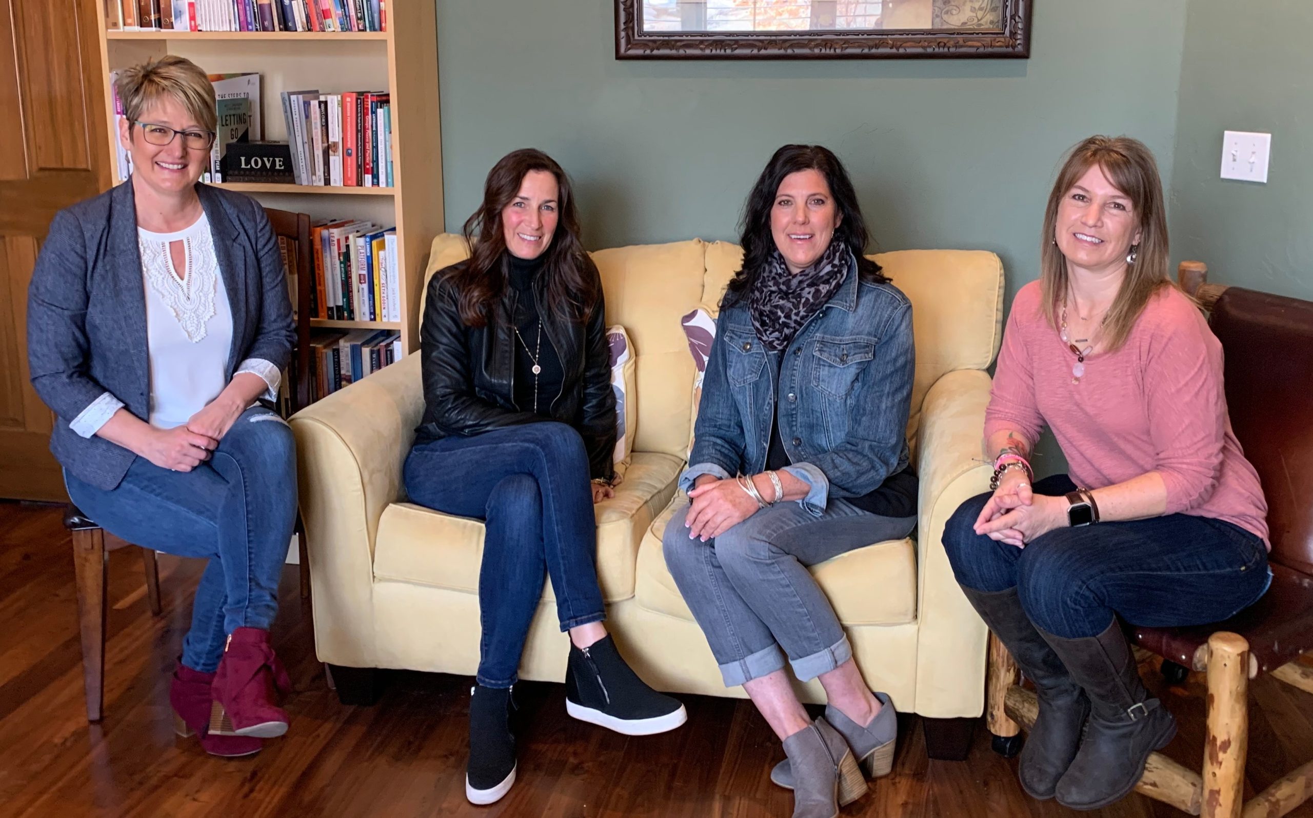 Four women sitting together on armchairs and a sofa in a room lined with bookshelves.
