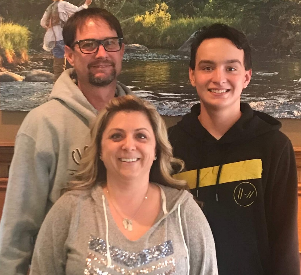 A family of three — two parents and a teenage son — smiling for a photograph beside a river.