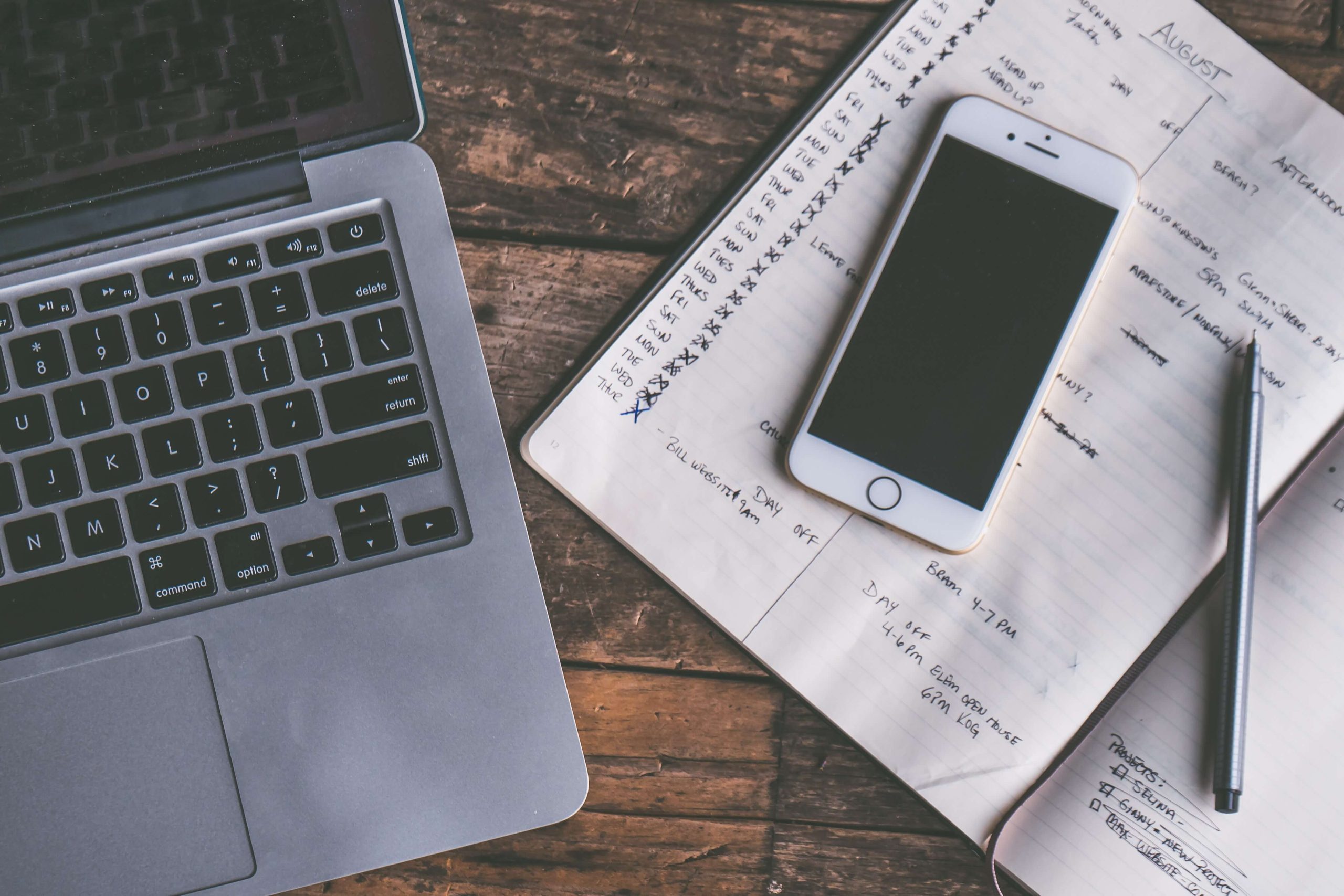 An open laptop, a phone and a pen resting on a handwritten notebook page on a wooden desk.