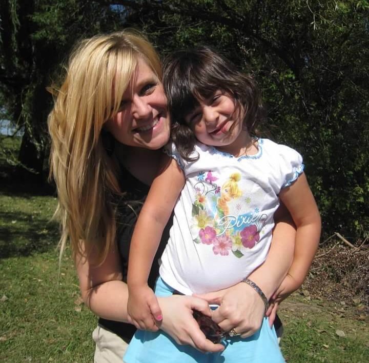 A woman with long blonde hair holding a smiling young girl in a floral T-shirt in a garden.