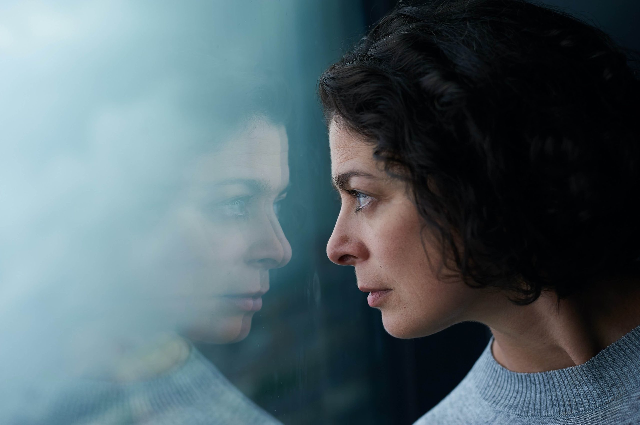 A woman resting her forehead against a window, her face reflected back at her in the glass.