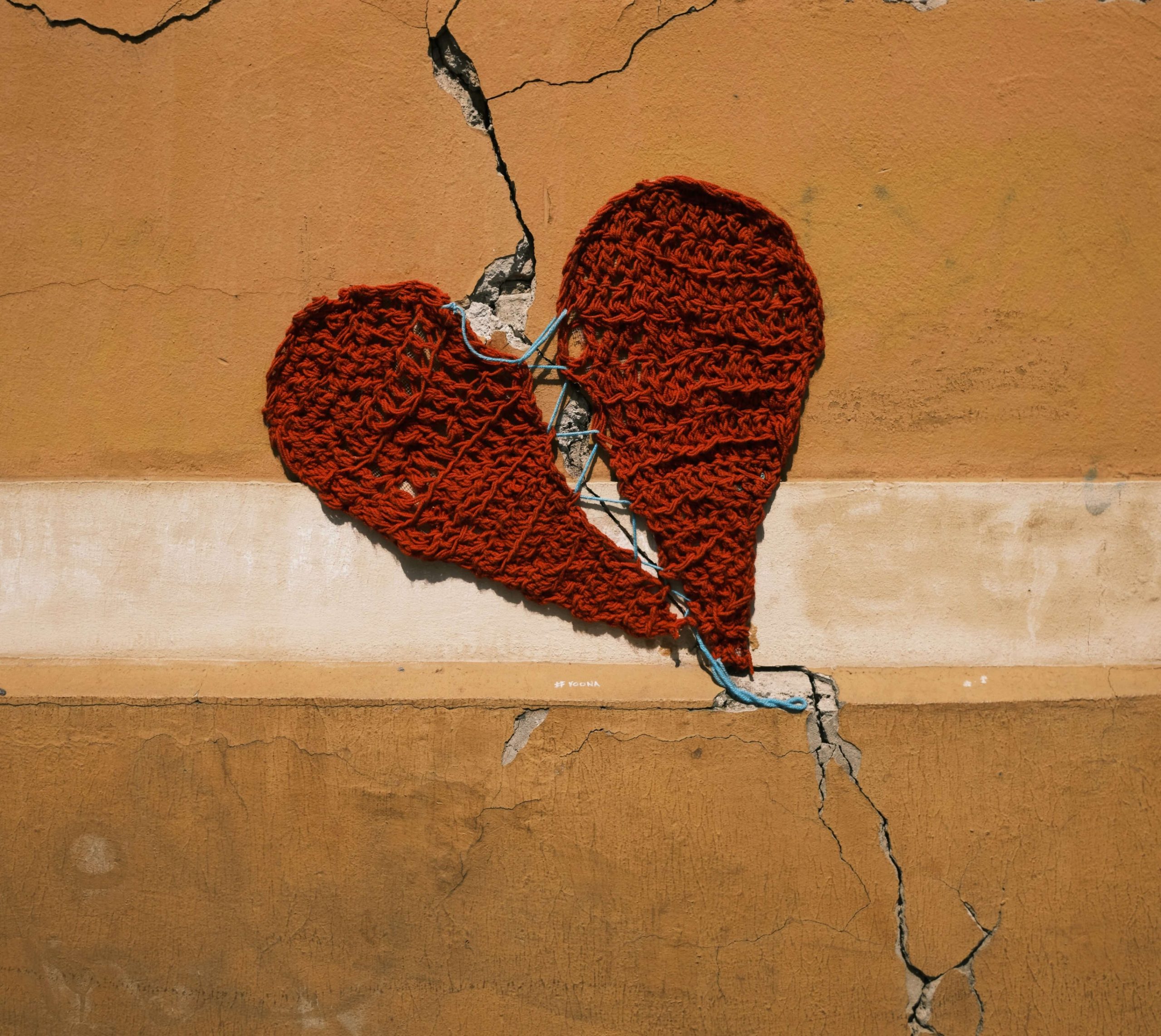 A crocheted red heart mounted over a crack running down a rendered wall.