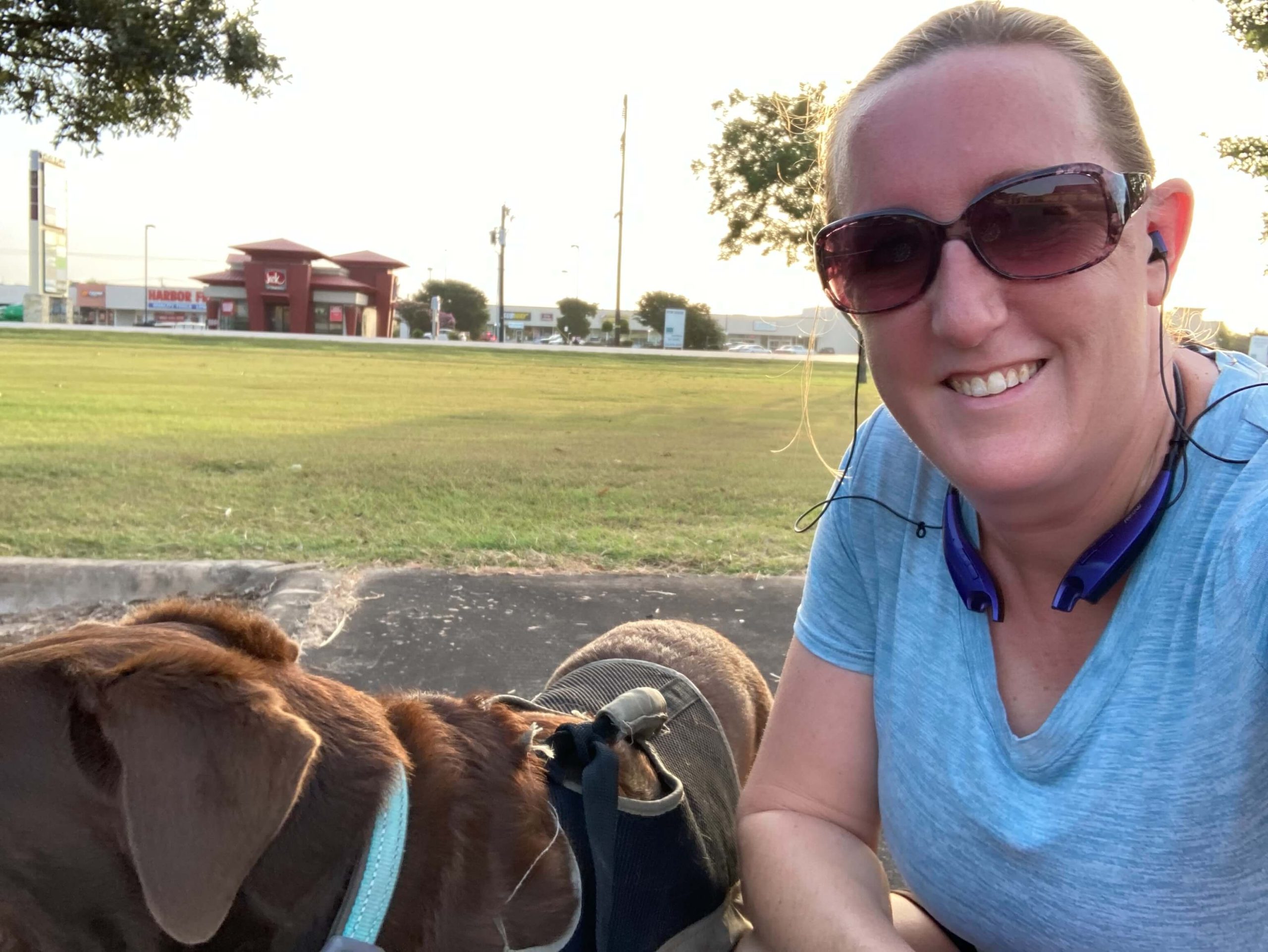 A woman in sunglasses smiling beside her chocolate labrador in a park.