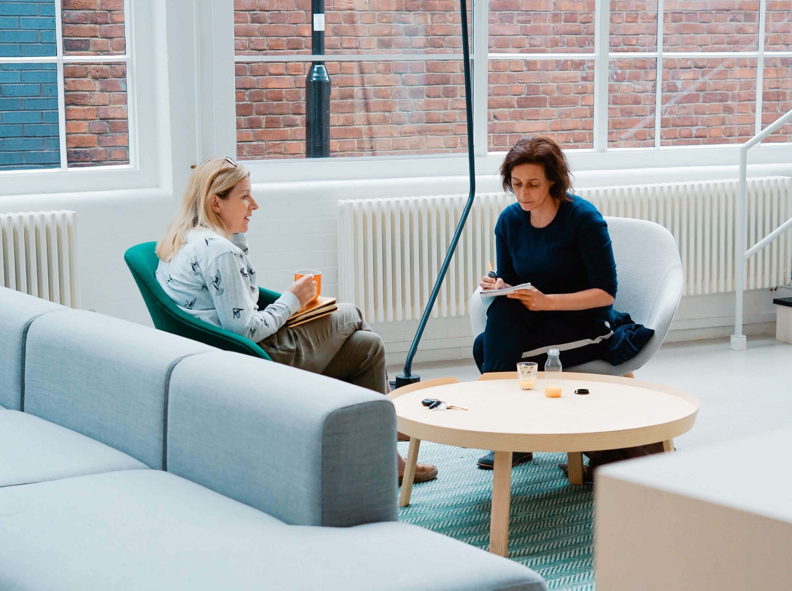 Two people sitting apart in a bright office lounge, one taking notes.