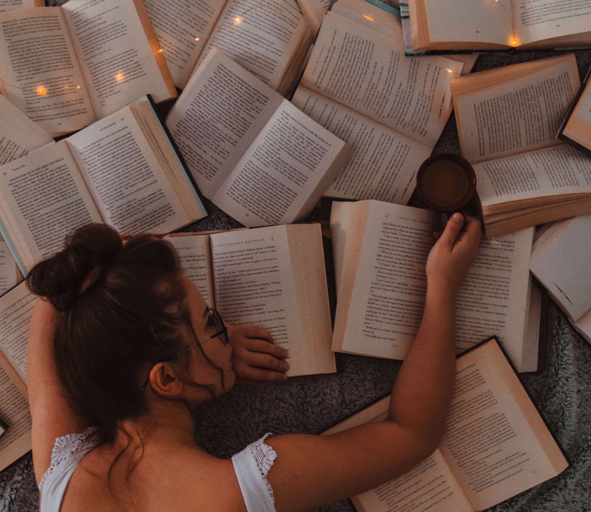 A young woman lying on the floor surrounded by dozens of open books, reaching for one.