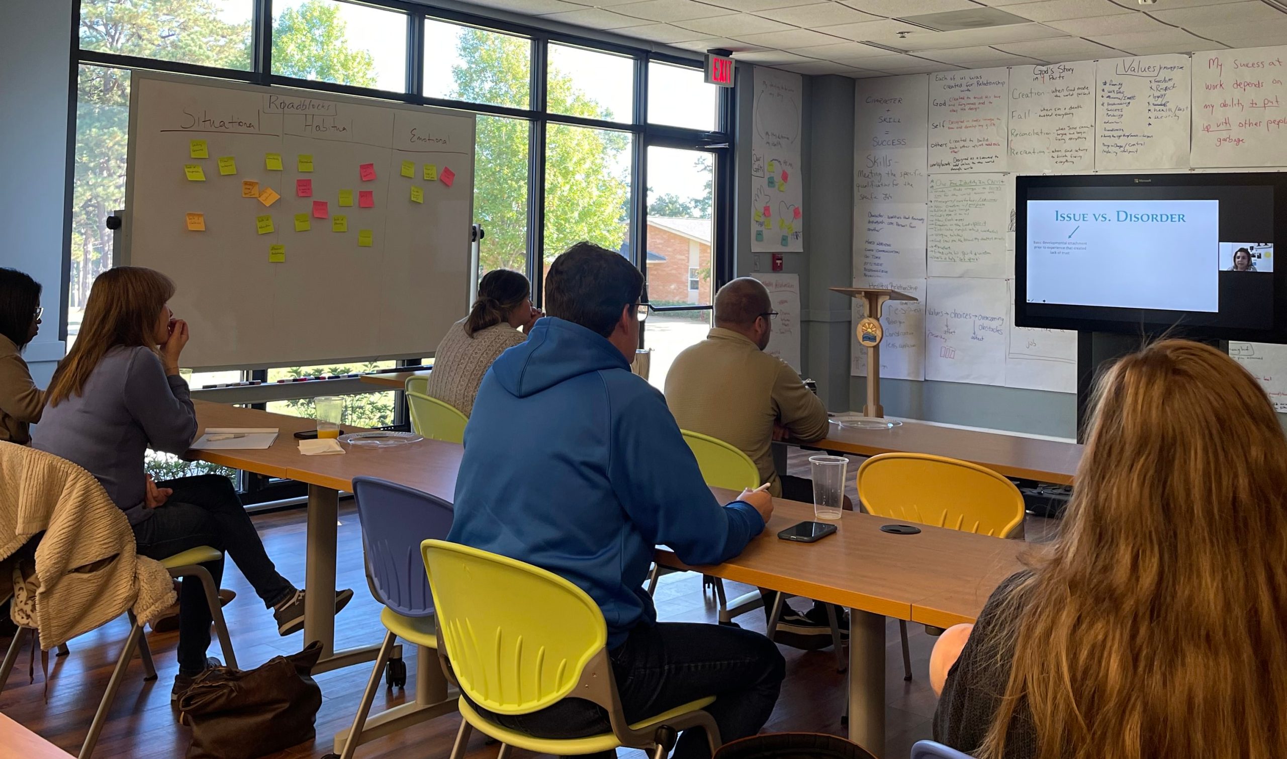 People seated around tables in a meeting room, facing a whiteboard covered in sticky notes.