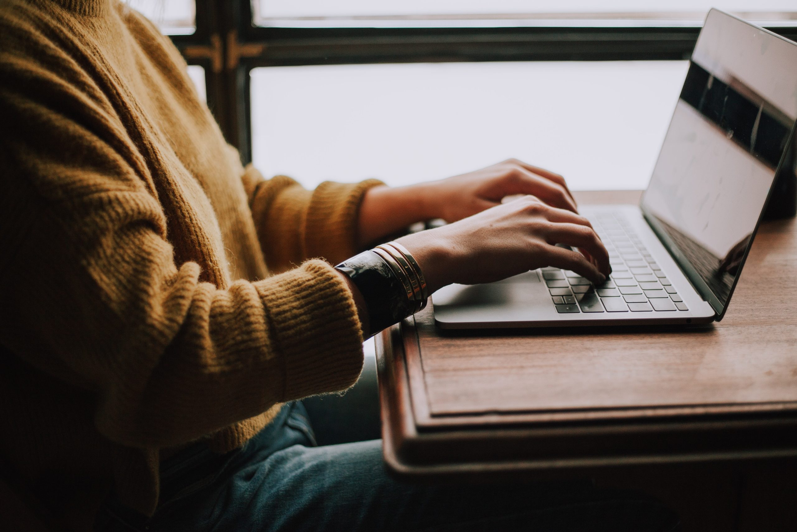 A person in a mustard jumper typing at a laptop by a window.