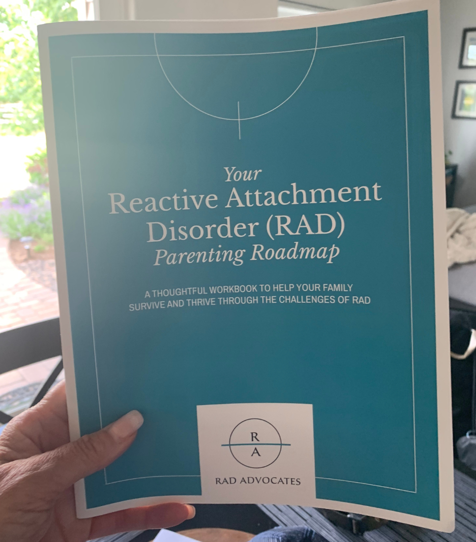 A hand holding a teal workbook titled "Your Reactive Attachment Disorder (RAD) Parenting Roadmap — a thoughtful workbook to help your family survive and thrive through the challenges of RAD", published by RAD Advocates.