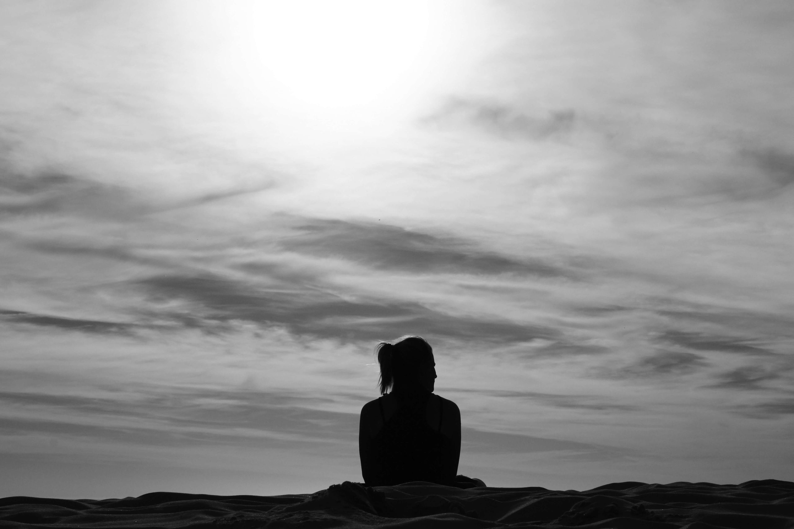 A person sitting alone on a shoreline in black and white, facing a bright, overcast sea.