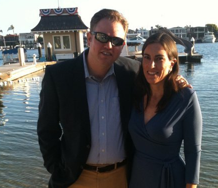 A man in sunglasses and a dark blazer standing arm in arm with a woman in a navy dress on a marina dock.