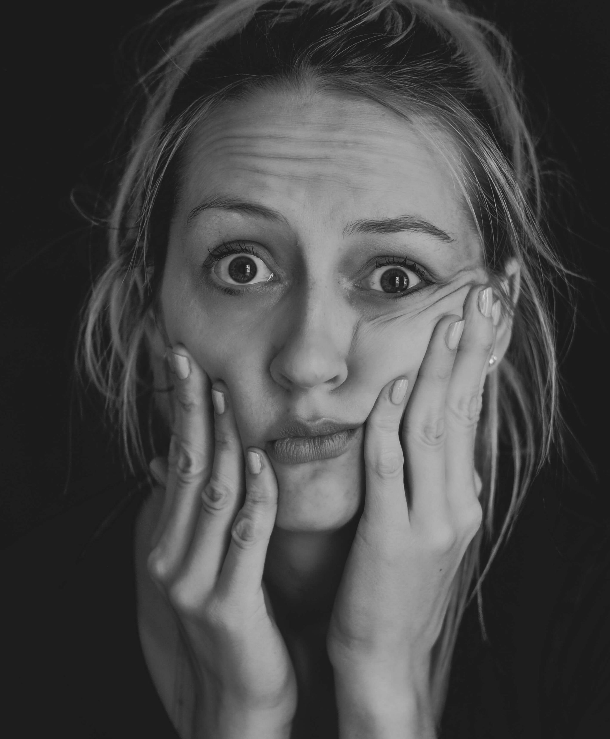 A black and white portrait of a woman holding her face in both hands, eyes wide.