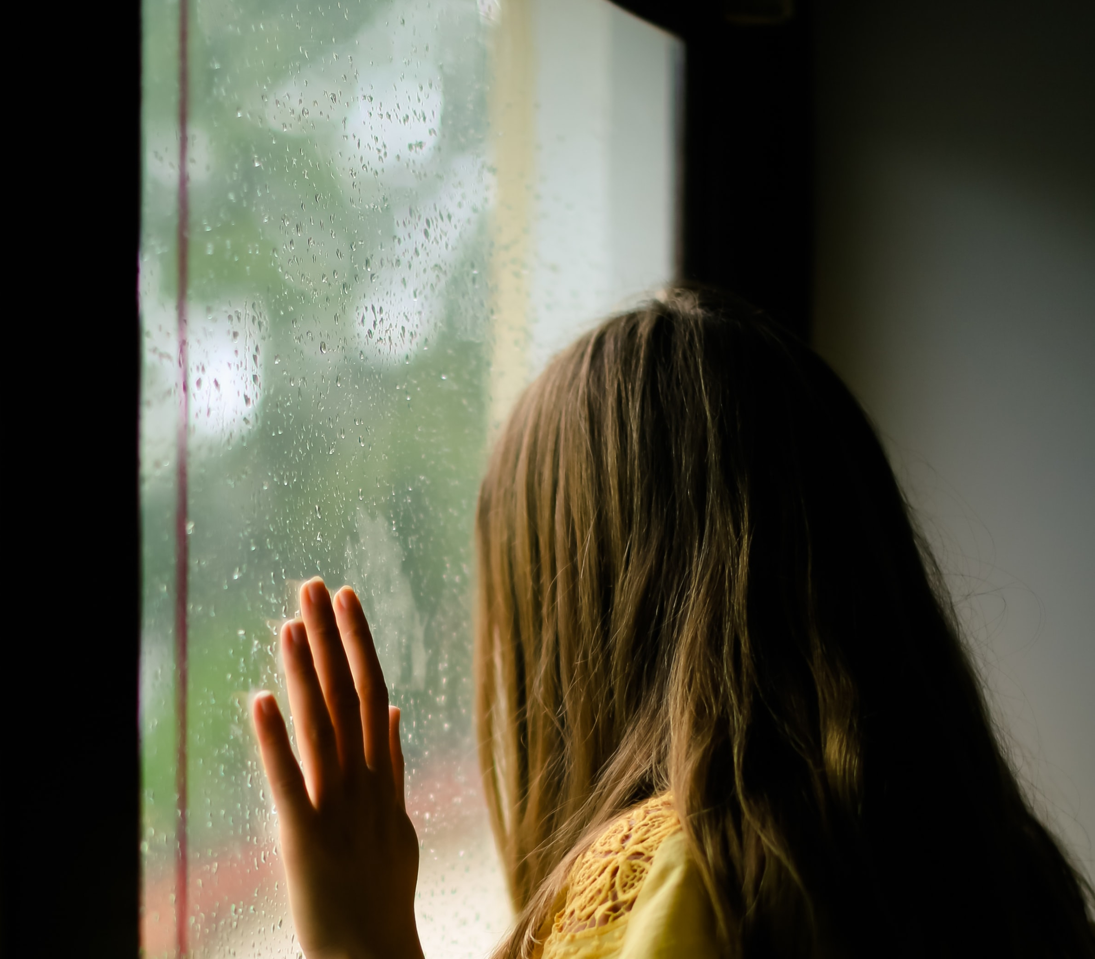 A girl resting her hand against a rain-streaked window, looking out.