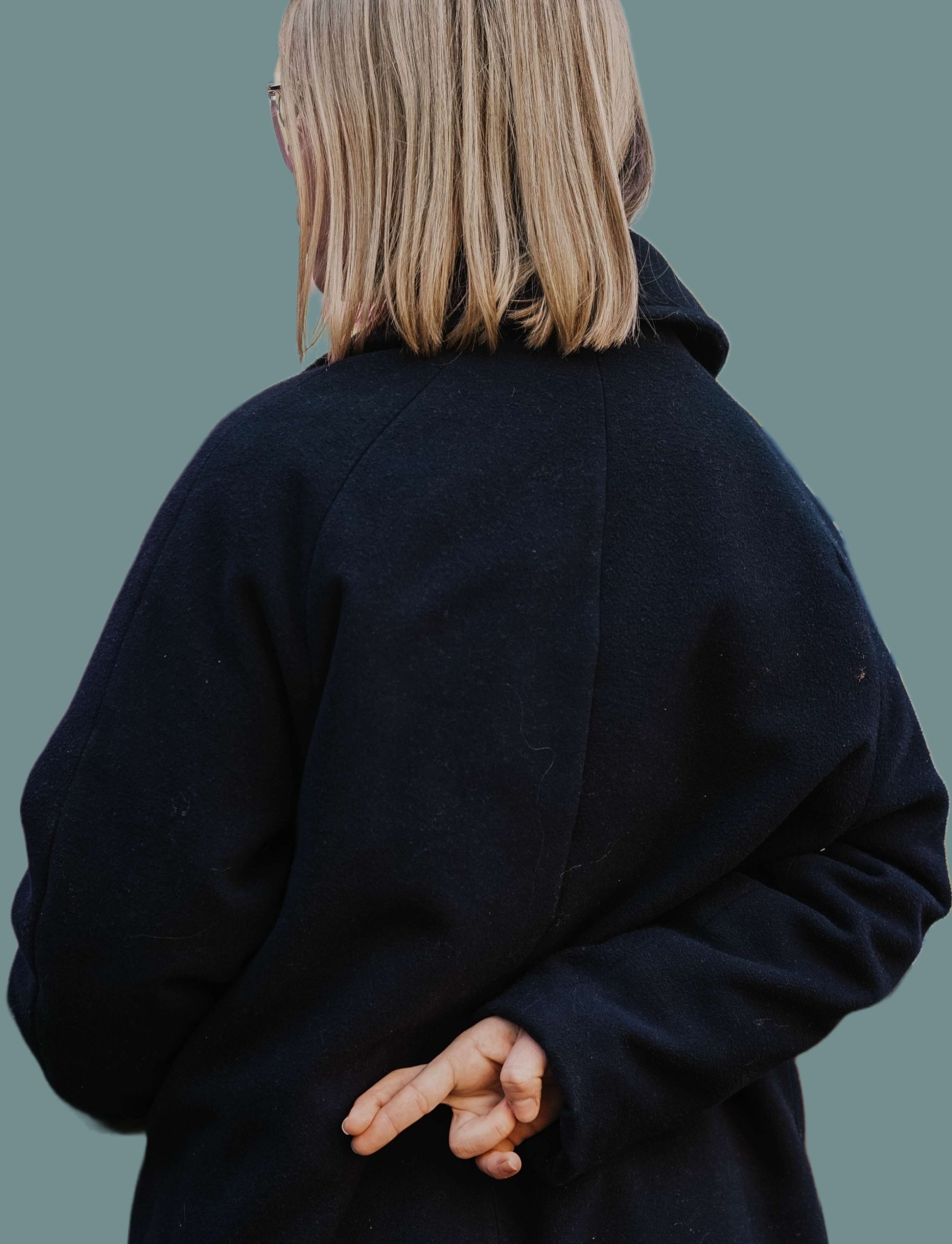A person in a dark coat photographed from behind, holding their fingers crossed behind their back.