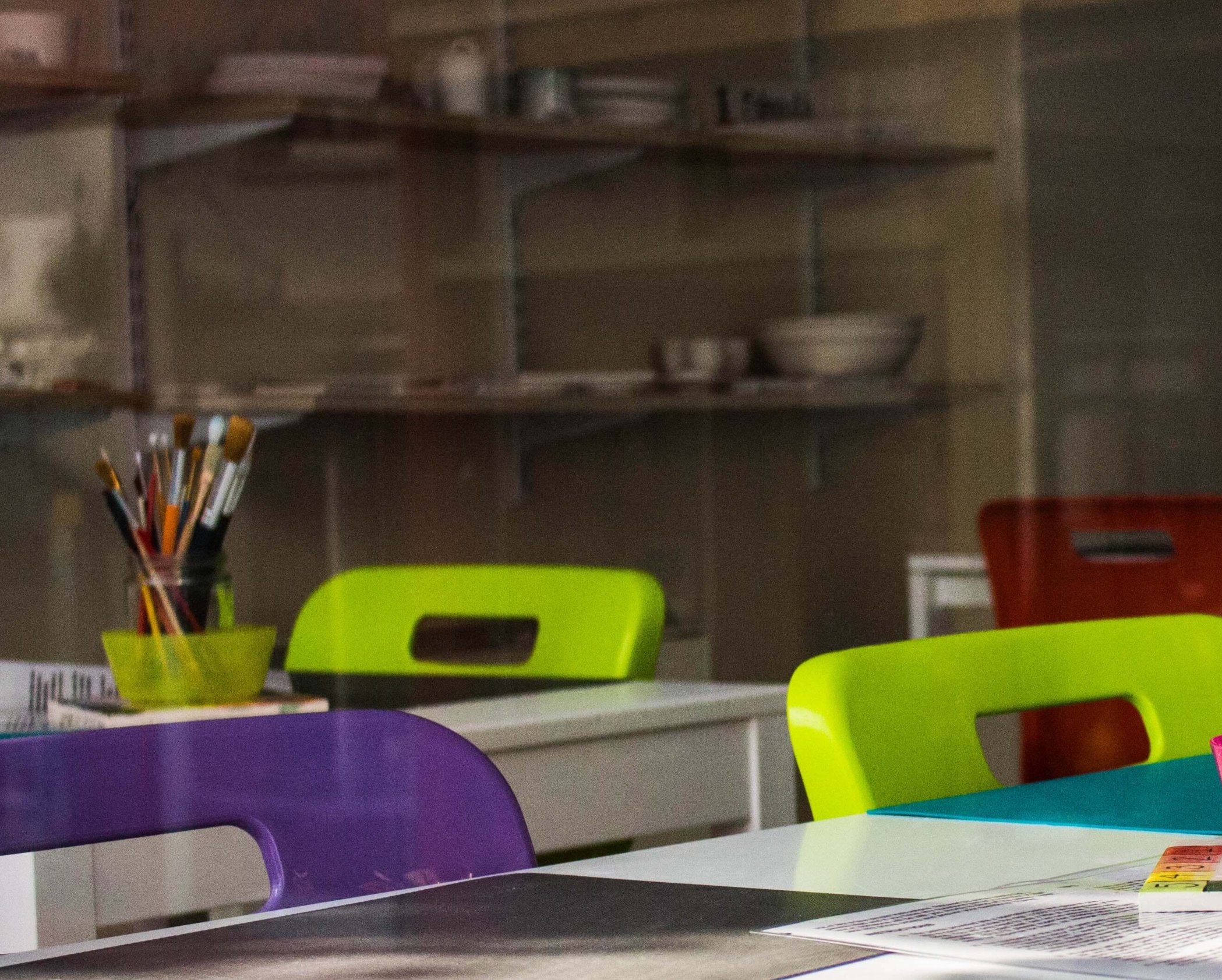 An empty classroom with brightly coloured chairs and pots of paintbrushes on the desks.