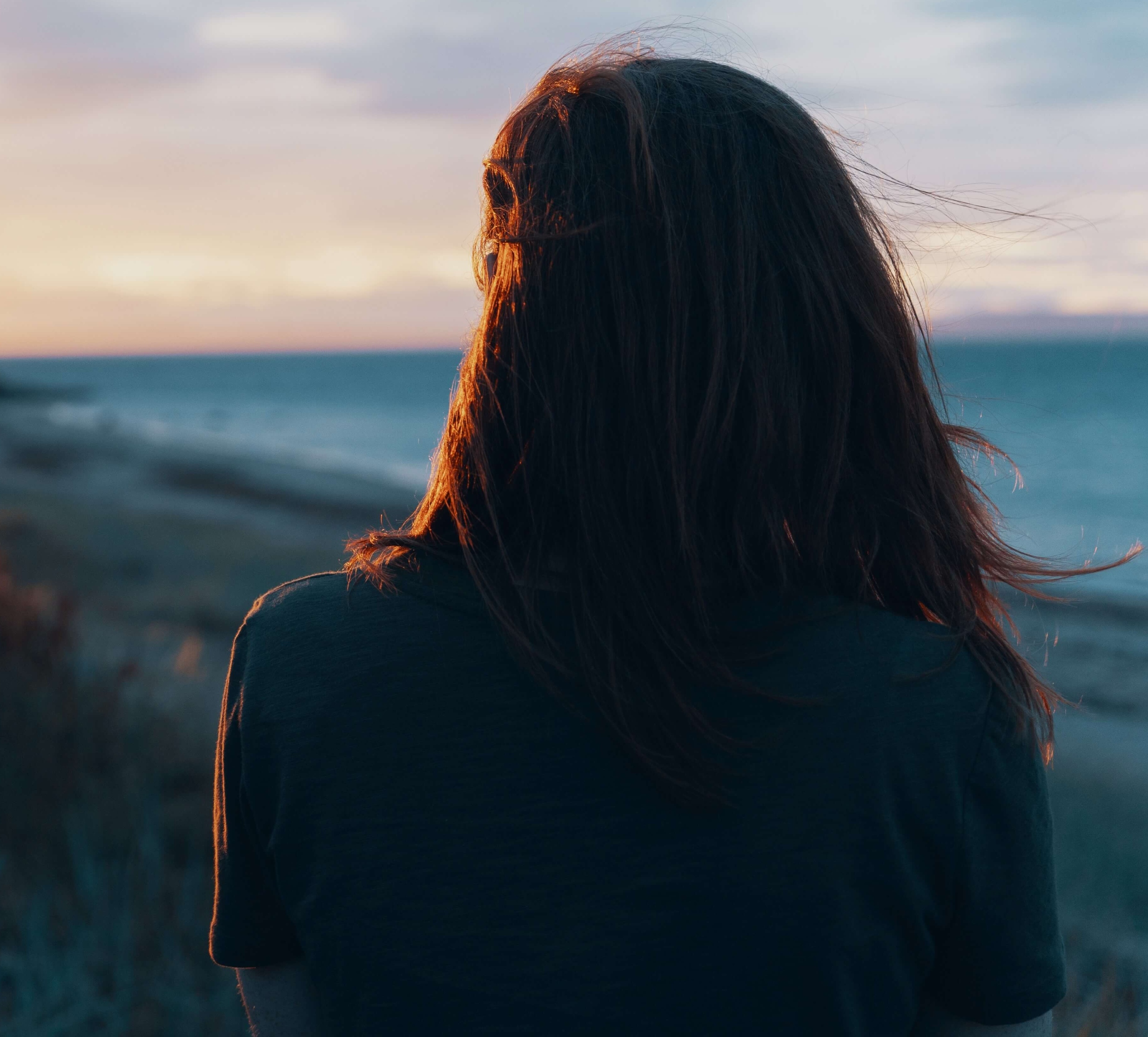A woman with long dark hair seen from behind, looking out over a beach at dusk.